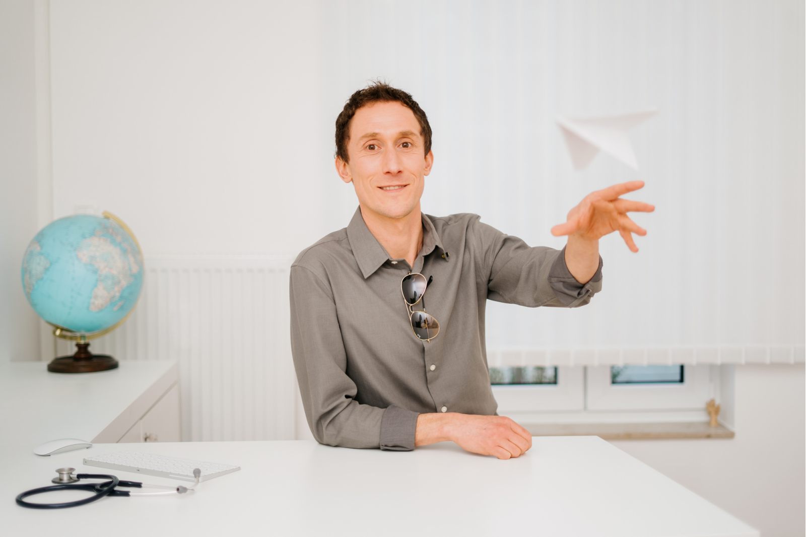 Ein Mann sitzt an einem Schreibtisch in einem hellen Büro und wirft spielerisch ein Papierflugzeug. Auf dem Schreibtisch stehen ein Globus, ein Stethoskop und eine Tastatur, und er hat eine Sonnenbrille an seinem Hemd hängen.