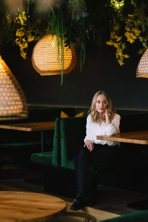 Eine Frau mit blondem Haar, weißer Bluse und schwarzer Hose, sitzt allein auf einem grünen Samttisch in einem Restaurant mit Holztischen und hängenden, geflochtenen Hängelampen, umgeben von Grünzeug.