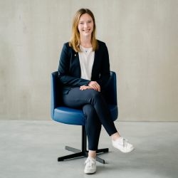 Eine junge Frau mit glattem, hellbraunem Haar lächelt, während sie auf einem modernen blauen Stuhl sitzt. Sie trägt einen marineblauen Blazer, ein weißes Hemd, eine marineblaue Hose und weiße Turnschuhe, wobei sie ihre Hände auf die gekreuzten Beine legt. Der Hintergrund ist schlicht und hellgrau.