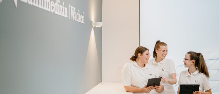 Drei Frauen in weißen Kitteln stehen lächelnd und redend in einer hellen, modernen Zahnklinik zusammen. Zwei halten Klemmbretter, eine hält ein Tablet. An der Wand steht der Text Zahnmedizin Wiesach.