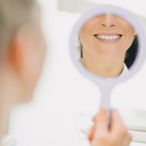 Eine Person hält einen runden Handspiegel und lächelt ihr Spiegelbild an. Der Fokus liegt auf ihren hellen, gesunden Zähnen, während ihr Gesicht im Vordergrund teilweise verschwommen ist. Der Hintergrund ist eine helle, saubere Umgebung.