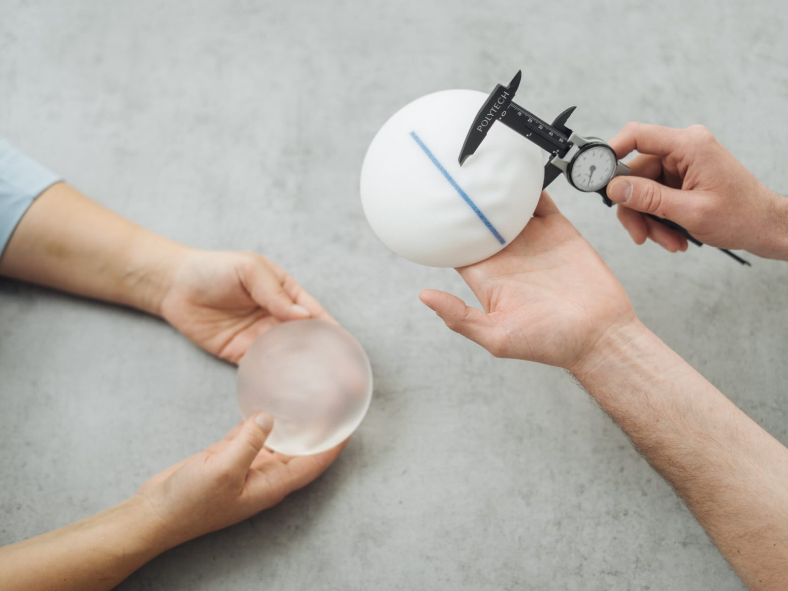 Zwei Personen halten Silikon-Brustimplantate; eine misst mit einem Messschieber ein weißes, strukturiertes Implantat, während die andere ein glattes, transparentes Implantat hält. Der Hintergrund ist eine hellgraue Fläche.