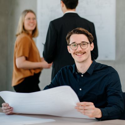 Ein lächelnder Mann mit Brille hält ein großes Blatt Papier an einem Tisch, während sich im Hintergrund eine Frau und ein Mann in Geschäftskleidung in einer modernen Büroumgebung unterhalten.
