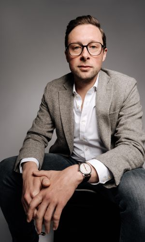 Ein Mann mit Brille, grauem Blazer, weißem Hemd und Jeans sitzt auf einem Hocker, hat die Hände gefaltet und blickt selbstbewusst in die Kamera vor einem schlichten grauen Hintergrund.