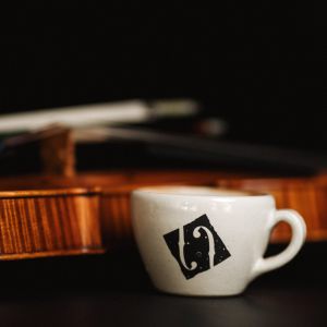 Eine kleine weiße Kaffeetasse mit schwarzem Muster steht vor einer Geige und einem Bogen, die beide im Hintergrund auf einer dunklen Fläche verschwimmen.
