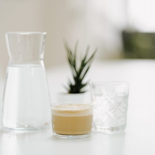 Eine Glaskaraffe mit Wasser, ein leeres strukturiertes Glas und eine Tasse Kaffee stehen auf einem weißen Tisch mit einer kleinen Topfpflanze, die im Hintergrund verschwimmt.