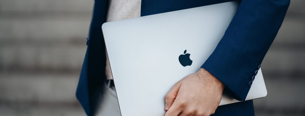 Eine Person, die einen blauen Blazer und eine helle Hose trägt, hält ein geschlossenes silbernes MacBook unter dem Arm und steht vor verschwommenen grauen Stufen.