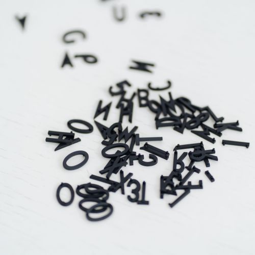Ein Stapel verstreuter schwarzer Plastikbuchstaben auf einer weißen Fläche, wobei einige Buchstaben im Vordergrund gruppiert und andere im Hintergrund verstreut sind.