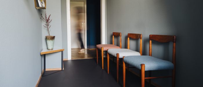 Ein Flur mit drei Holzstühlen mit farbigen Sitzen an einer grauen Wand, ein kleiner Tisch mit einer Vase, Wandregale mit Büchern und eine verschwommene Person, die durch eine offene Tür im Hintergrund geht.