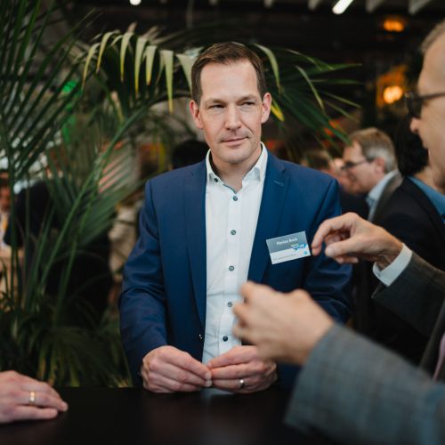 Ein Mann in einem blauen Anzug mit einem Namensschild steht an einem Tisch und hört zwei anderen Männern in Anzügen bei einem Networking-Event zu. Im Hintergrund sind Grünpflanzen und mehrere Menschen zu sehen.