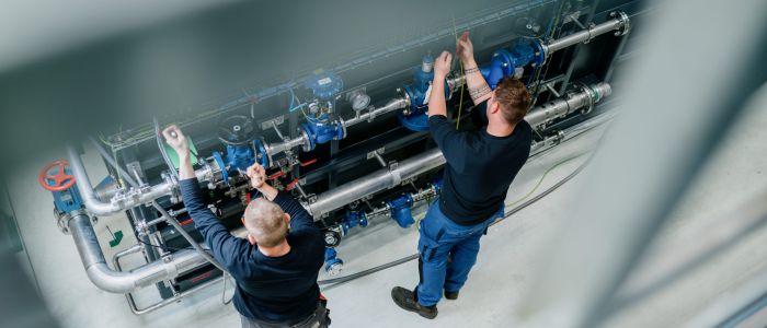 Zwei Arbeiter in Uniform überprüfen und justieren Industrierohre und Maschinen in einer sauberen, modernen Anlage, von oben gesehen.