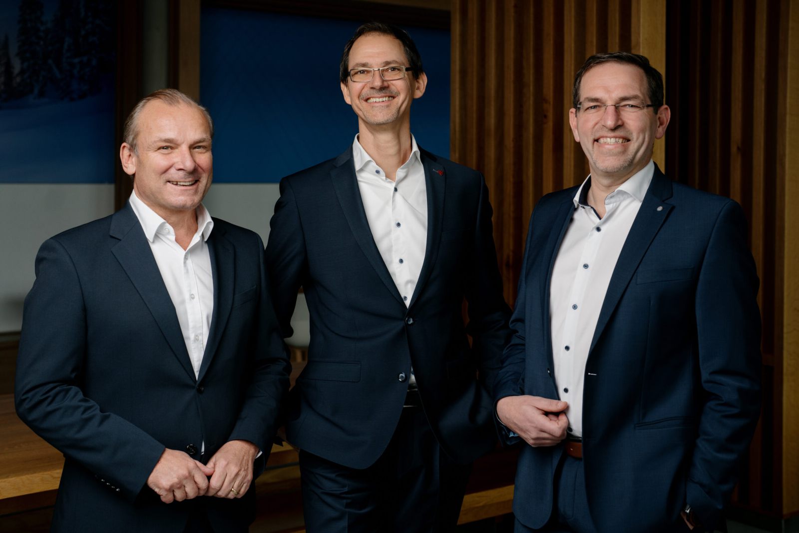 Drei Männer in dunklen Anzügen und weißen Hemden stehen in einem Haus und lächeln in die Kamera. Sie scheinen sich in einer professionellen oder geschäftlichen Umgebung zu befinden, mit Holztafeln und einem blauen Hintergrund hinter ihnen.