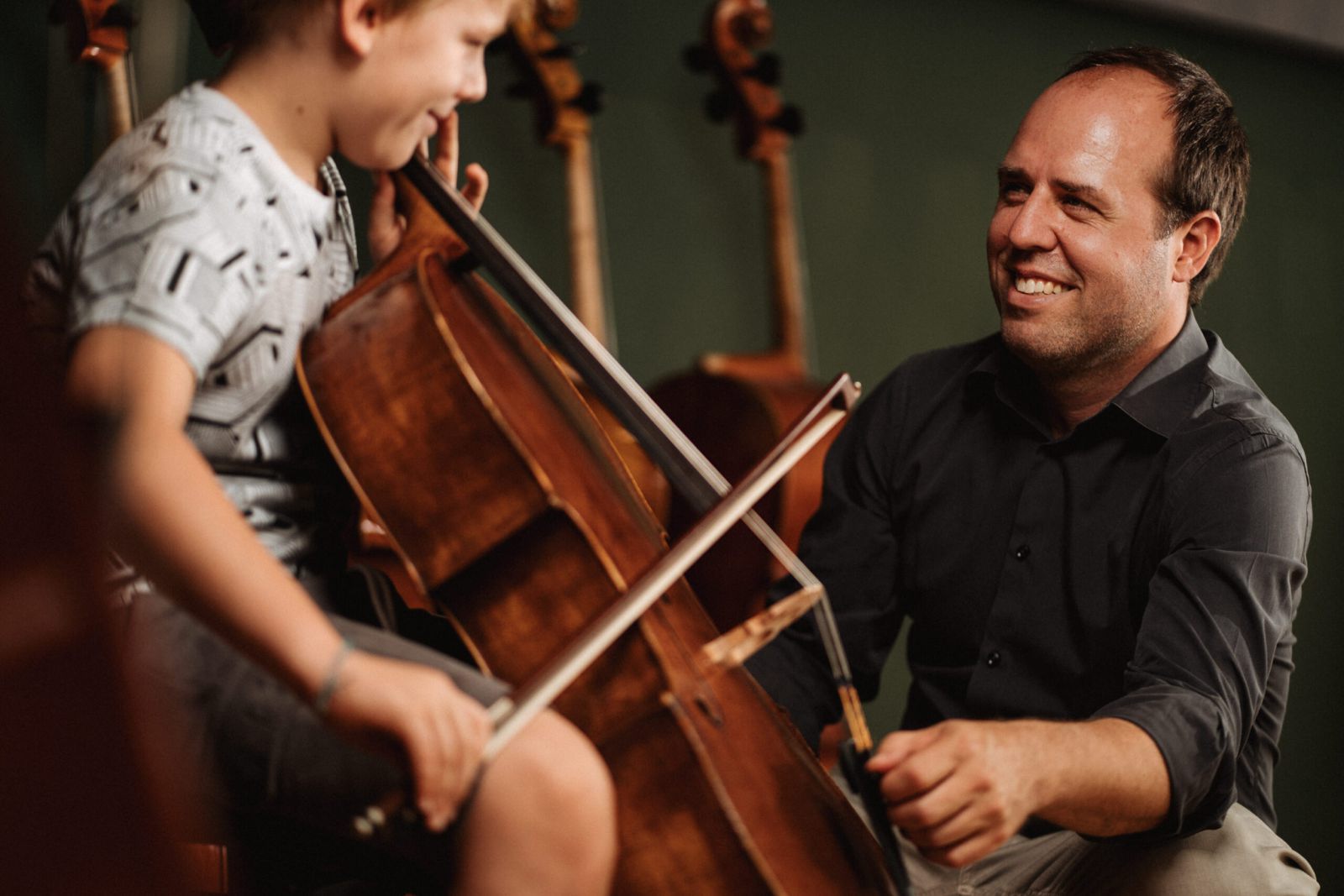 Ein lächelnder Mann hilft einem kleinen Jungen beim Cellospiel, indem er den Bogen über die Saiten führt, während im Hintergrund Celli zu sehen sind.