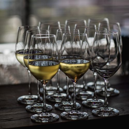 Eine Gruppe von Weingläsern auf einer hölzernen Oberfläche, wobei mehrere Gläser mit Weißwein gefüllt und andere leer sind, vor einem verschwommenen Hintergrund mit weicher Beleuchtung.