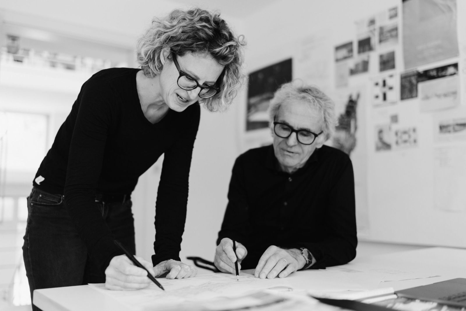 Eine Frau und ein älterer Mann, beide mit Brille, arbeiten an einem Schreibtisch in einem hellen Büro an Skizzen, während hinter ihnen Pläne und Bilder an der Wand hängen. Das Bild ist schwarz-weiß.