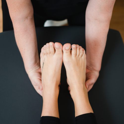 Eine Person erhält eine Fußmassage, während sie auf einer schwarzen Massageliege liegt. Der Fokus liegt auf ihren nackten Füßen und den Händen des Masseurs, die sie sanft halten. Im Hintergrund ist ein Holzboden zu sehen.
