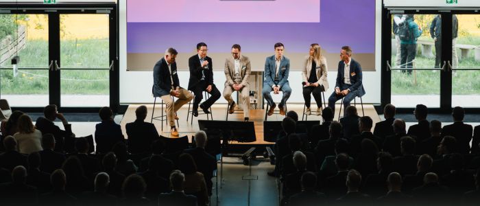 Sieben Personen sitzen auf einer Bühne vor einem Publikum und nehmen an einer Podiumsdiskussion teil. Große Fenster hinter ihnen zeigen Grünflächen und zwei Personen, die draußen stehen. Die Umgebung scheint eine Konferenz oder ein Seminar zu sein.