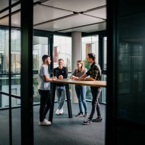 Vier junge Erwachsene stehen um einen hohen Tisch in einem modernen Büro mit Glaswänden, unterhalten sich und lächeln. Der Raum ist von natürlichem Licht durchflutet, und vor den Fenstern sind die Gebäude der Stadt zu sehen.