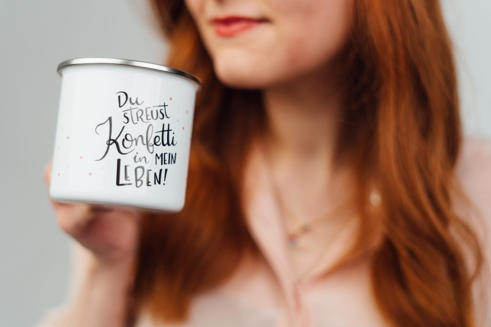 Eine Frau mit langen roten Haaren hält einen weißen Becher mit der Aufschrift "Du streust Konfetti in mein Leben! Der Fokus liegt auf dem Becher, und ihr Gesicht ist teilweise unscharf.