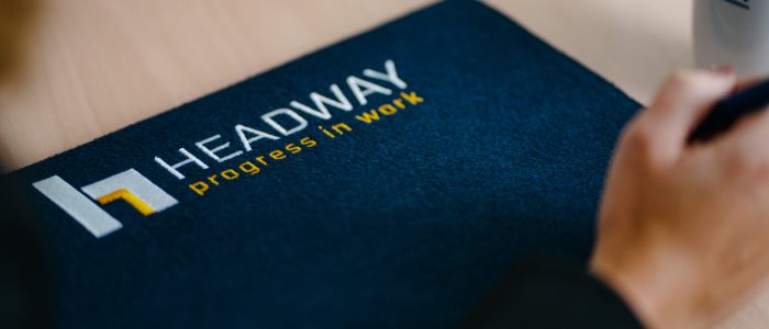 Nahaufnahme einer dunkelblauen Mappe mit dem Logo HEADWAY progress in work in weißer und gelber Schrift. Im Hintergrund sind teilweise eine Personenhand und eine Tasse mit der Aufschrift Headway zu sehen.