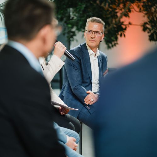Ein Mann in einem blauen Anzug sitzt in einer Podiumsdiskussion und hört aufmerksam einem anderen Diskussionsteilnehmer zu, der ein Mikrofon hält. Zwei weitere unscharfe Figuren befinden sich im Vordergrund, und im Hintergrund ist Grün zu sehen.