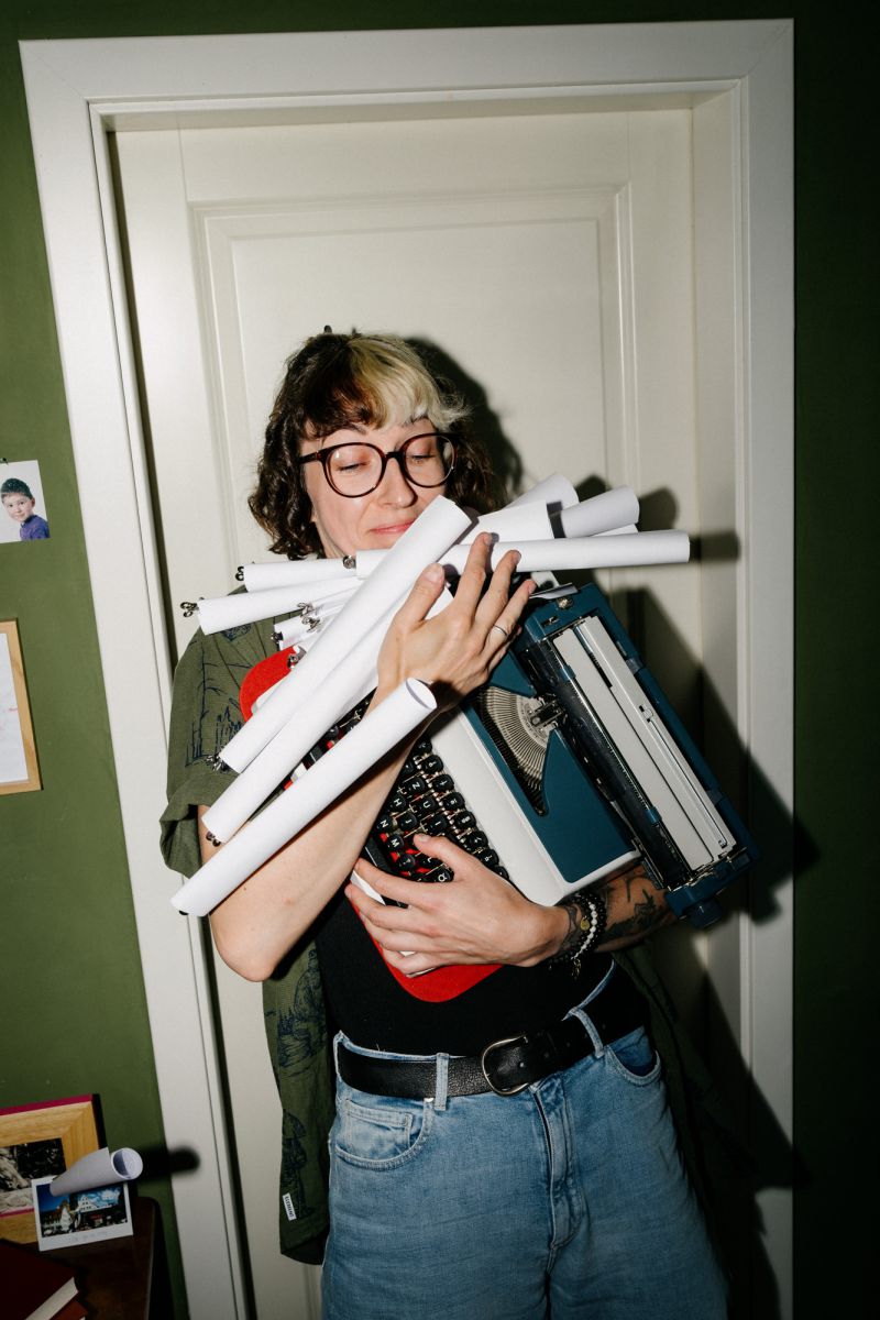 Eine Person mit Brille und lockigem Haar steht vor einer Tür, lächelt und hält mehrere zusammengerollte Papiere und eine Schreibmaschine im Arm. Sie trägt ein grünes Hemd und eine blaue Jeans.