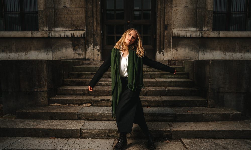 Eine Frau mit langen blonden Haaren steht auf steinernen Stufen vor einem großen, dunklen Gebäude. Sie trägt einen langen Mantel, Stiefel und einen grünen Schal, hat die Arme leicht ausgestreckt und den Kopf geneigt.