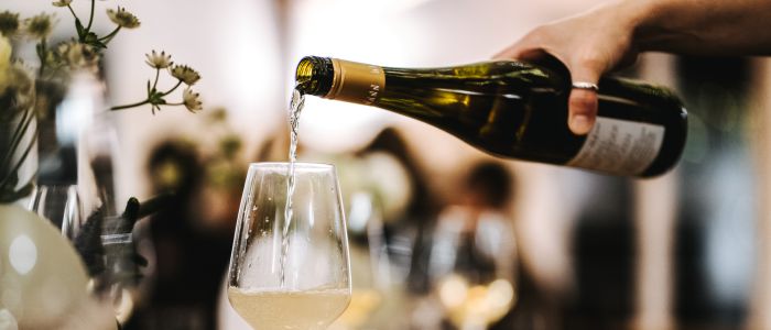 Eine Hand gießt Weißwein aus einer Flasche in ein Weinglas auf einem gedeckten Esstisch, mit verschwommenen Blumen und Geschirr im Hintergrund.