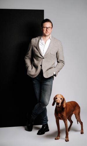 Ein Mann in einem hellgrauen Blazer, weißem Hemd und Jeans steht mit den Händen in den Taschen an eine schwarze Tafel gelehnt. Ein brauner Hund steht neben ihm. Der Hintergrund ist schlicht und hell.