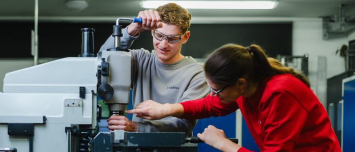 Zwei Personen bedienen eine Fräsmaschine in einer Werkstatt. Einer stellt die Bedienelemente ein, während der andere ein Werkstück führt, beide konzentriert und mit Schutzbrille. Im Hintergrund sind blaue Schränke und Maschinen zu sehen.