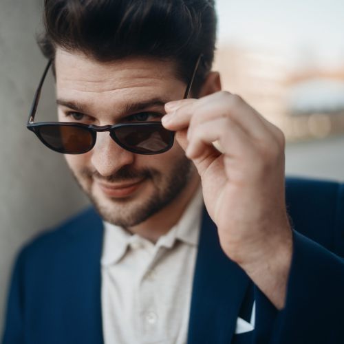 Ein Mann mit dunklem Haar und Gesichtsbehaarung trägt einen blauen Blazer und ein weißes Hemd und blickt mit einem leichten Lächeln über seine Sonnenbrille, während er im Freien vor einem unscharfen Hintergrund steht.