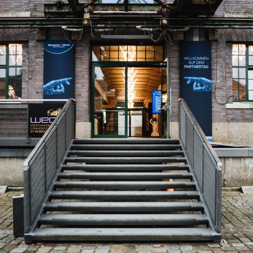 Eine graue Metalltreppe führt zum Eingang eines Backsteingebäudes mit Glastüren. Blaue Banner mit deutschem Text flankieren die Türen, und im Vordergrund ist ein kopfsteingepflasterter Weg zu sehen.