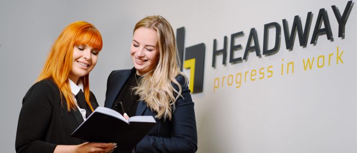 Zwei Frauen in Geschäftskleidung stehen lächelnd und mit Blick auf ein aufgeschlagenes Notizbuch neben einer Wand mit dem Logo HEADWAY progress in work.