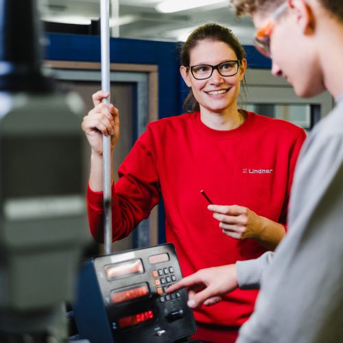 Zwei Personen arbeiten gemeinsam an einem Bedienfeld in einem technischen oder industriellen Umfeld. Eine Person in einem roten Sweatshirt lächelt und hält ein Werkzeug, während die andere die Einstellungen an der Maschine vornimmt.