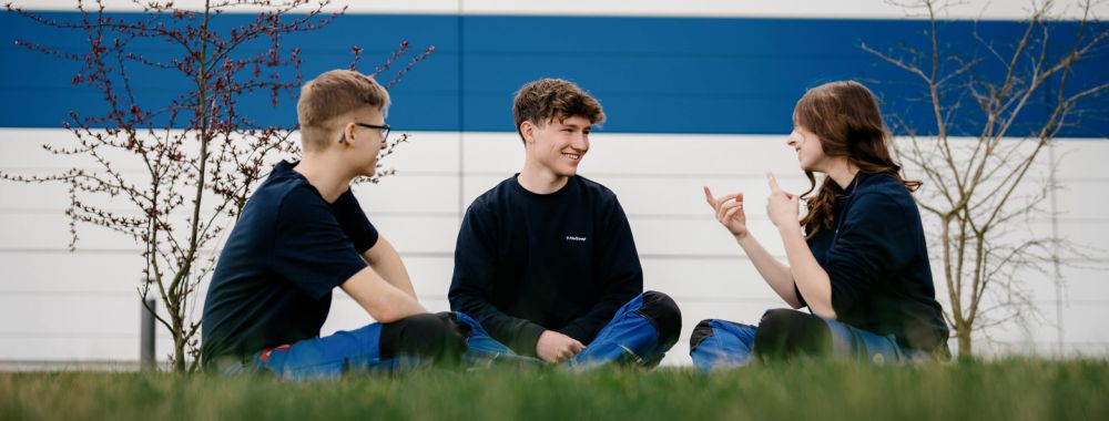 Drei junge Menschen sitzen im Schneidersitz im Gras, einander zugewandt und lächeln, im Hintergrund eine weiß-blau gestreifte Wand und kleine blattlose Bäume.