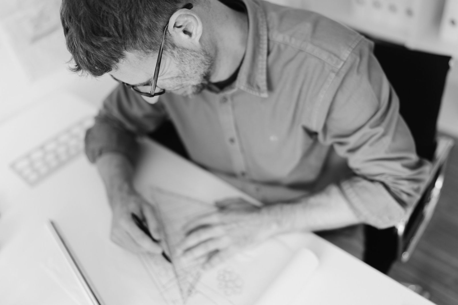 Schwarz-Weiß-Bild eines Mannes mit Brille, der an einem Schreibtisch sitzt und geometrische Formen auf einem großen Blatt Papier skizziert, möglicherweise bei der Arbeit an einem Architektur- oder Designprojekt.