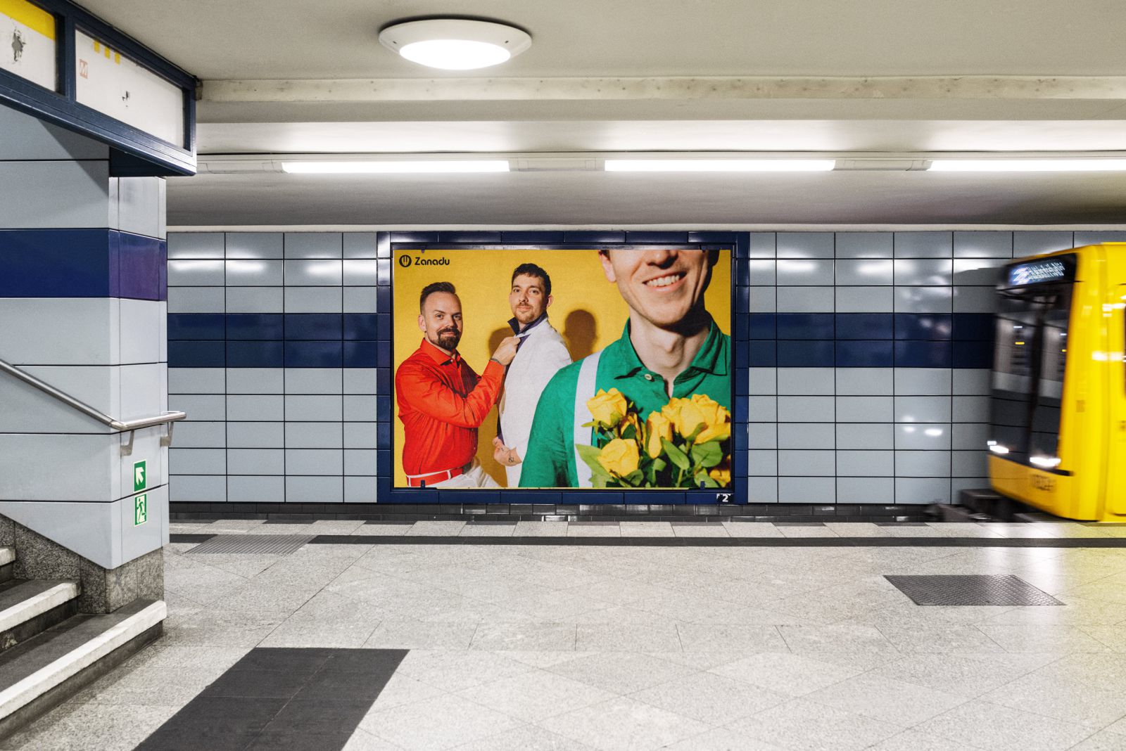 Auf einem U-Bahnsteig mit gekachelten Wänden ist eine große Werbung zu sehen, die zwei lächelnde Männer, einen in Rot und einen in Grau, hinter einer weiteren Person in einem grünen Hemd zeigt, die gelbe Blumen hält. Ein gelber Zug nähert sich von rechts.