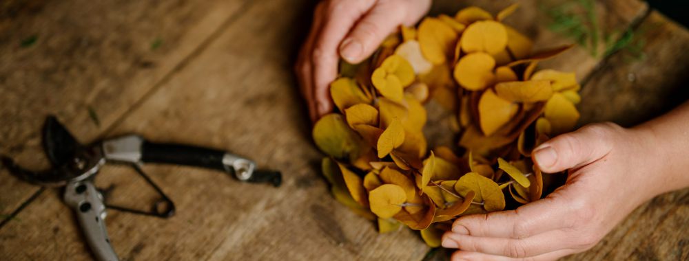 Die Hände einer Person formen einen Kranz aus gelben Herbstblättern auf einem rustikalen Holztisch, daneben liegen eine Gartenschere und eine Stecknadel mit rotem Kopf.