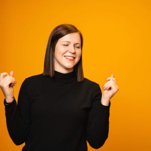 Eine Frau mit schwarzem Rollkragenpullover lächelt mit geschlossenen Augen und erhobenen Fingern fröhlich vor einem leuchtend orangefarbenen Hintergrund.