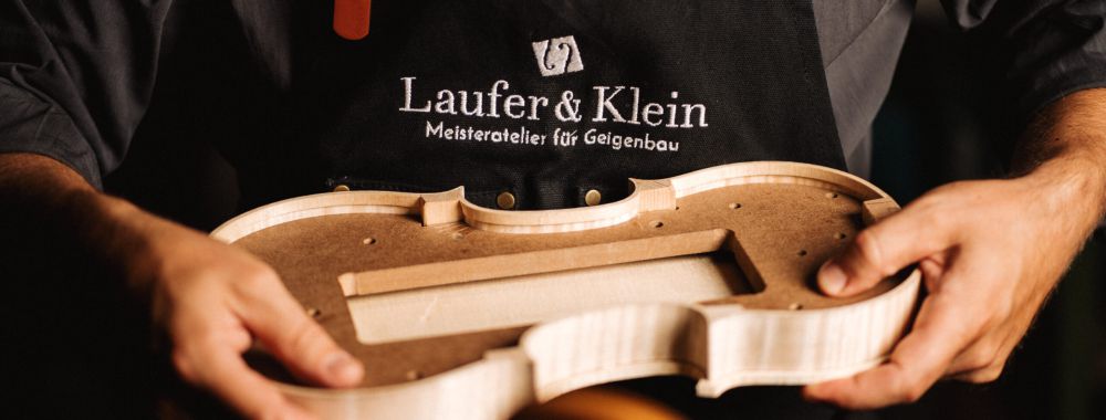 Eine Person, die eine schwarze Schürze mit der Aufschrift "Laufer & Klein Meisteratelier für Geigenbau" trägt, hält einen unfertigen Geigenkörper in einer Geigenbauwerkstatt.