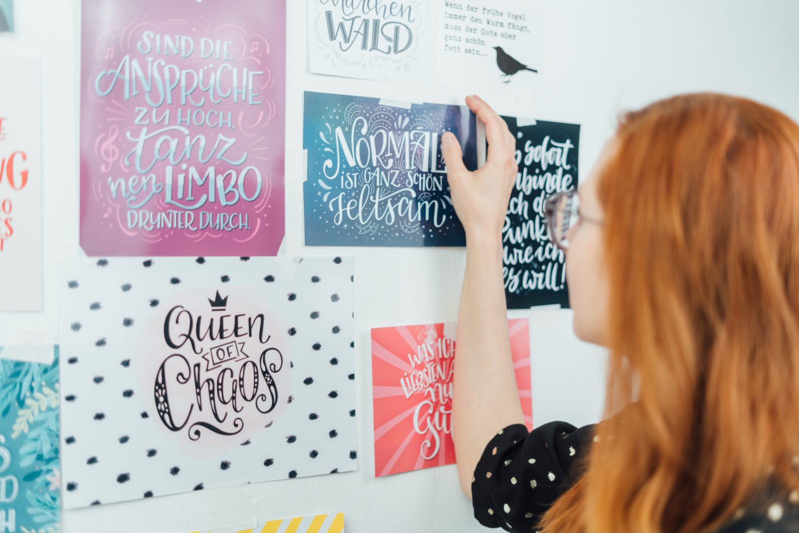 Eine Frau mit langen roten Haaren und Brille arrangiert bunte, handbeschriftete Motivationsposter an einer weißen Wand. Die Poster zeigen inspirierende deutsche Zitate mit künstlerischen, dekorativen Designs.