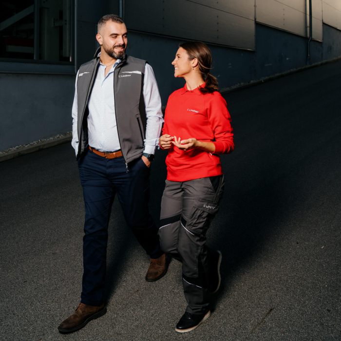 Ein Mann in einer grauen Weste und eine Frau in einem roten Sweatshirt gehen nebeneinander auf einer gepflasterten Straße neben einem großen Industriegebäude, unterhalten sich und lächeln.
