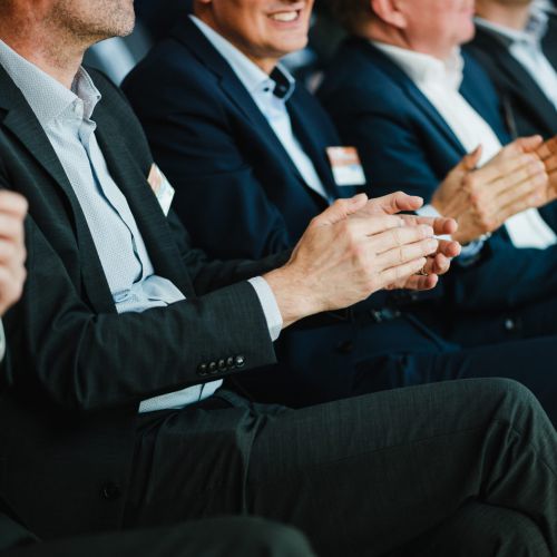 Eine Gruppe von Männern in Geschäftsanzügen sitzt in einer Reihe, lächelt und klatscht, wahrscheinlich bei einer offiziellen Veranstaltung oder Konferenz. Ihre Gesichter sind teilweise zu sehen, und an ihren Jacken sind Namensschilder befestigt.