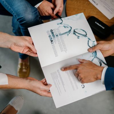 Zwei Personen, die an einem Tisch sitzen, diskutieren über ein gedrucktes Dokument mit Text und Bildern von Brillen, während eine andere Person auf einen Abschnitt des Papiers zeigt. Andere Papiere und ein Brillenetui liegen auf dem Tisch.