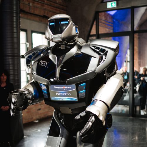 Ein großer humanoider Roboter mit einem glänzenden schwarz-silbernen Körper steht in einem Innenraum auf einem dunklen Boden, umgeben von Menschen und Ziegelwänden, beleuchtet von natürlichem und blauem Licht.