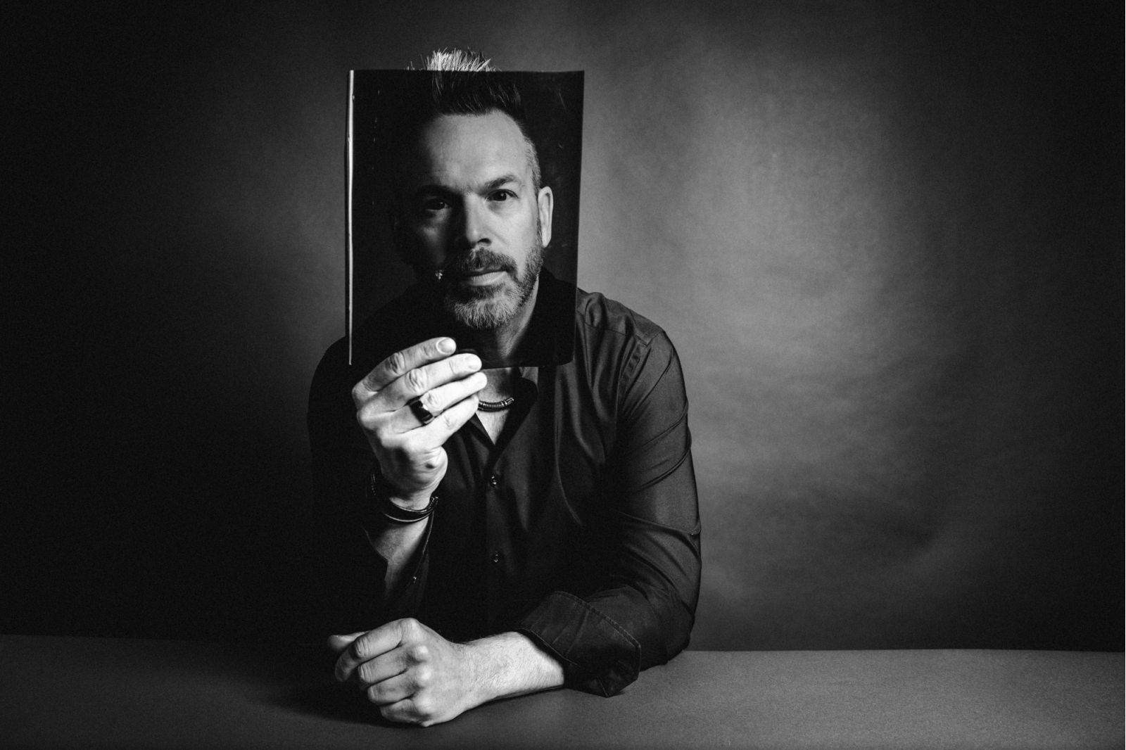 Ein Mann sitzt an einem Tisch und hält ein Schwarz-Weiß-Foto seines eigenen Gesichts vor sich, wobei sein echtes Gesicht mit dem Foto verschmilzt. Der Hintergrund ist dunkel und schafft eine dramatische und künstlerische Atmosphäre.