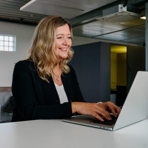 Eine Frau mit blonden Haaren, die einen schwarzen Blazer trägt, lächelt, während sie an einem Schreibtisch auf einem Laptop tippt. Im Hintergrund sitzen drei Personen auf einer Couch und unterhalten sich in einem modernen Büroraum.