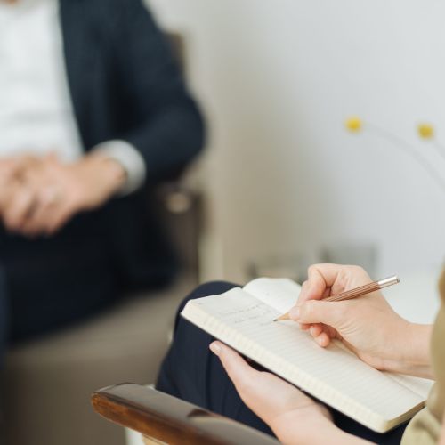 Eine Person sitzt mit gefalteten Händen im Hintergrund, während eine andere Person im Vordergrund Notizen in ein Notizbuch schreibt, was auf eine Therapie- oder Beratungssitzung schließen lässt.