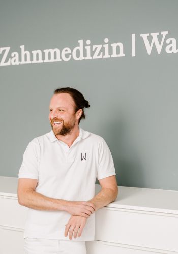 Ein Mann in einem weißen Polohemd steht lächelnd in einer modernen Zahnarztpraxis, hinter ihm steht Zahnmedizin Wackerl an der Wand.
