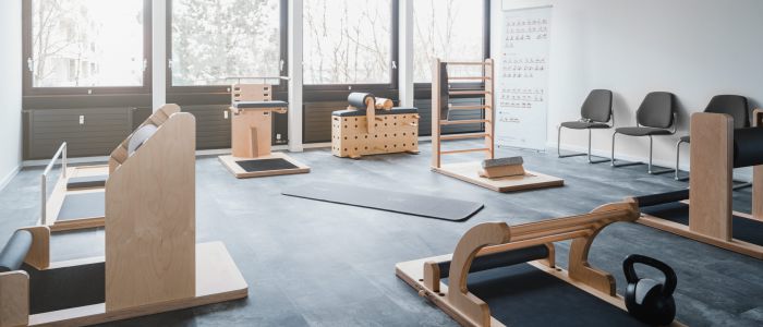 Ein heller, moderner Fitnessraum mit Fitnessgeräten aus Holz, einer schwarzen Kettlebell, großen Fenstern, die natürliches Licht hereinlassen, und einer Reihe grauer Stühle an der Wand.